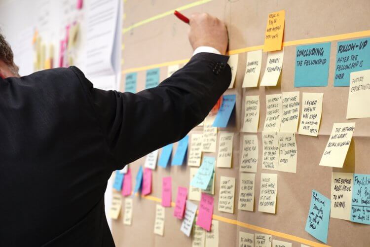 A man in a suit, a legal project manager, writes on a post-it note, which is stuck to a corkboard with dozens of other post-it notes