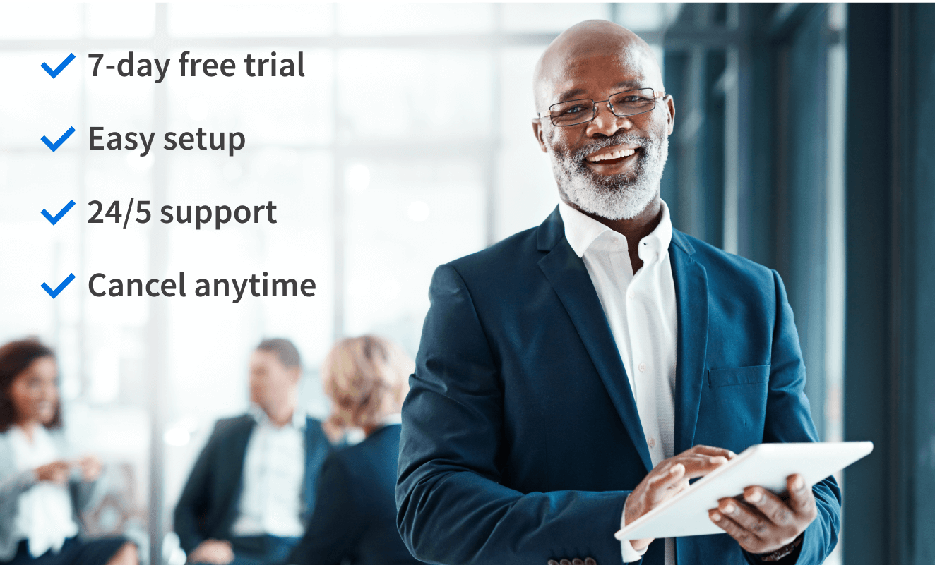 Man wearing a navy suit and white button-up shirt, standing in the foreground, holding a tablet and smiling. Three people in professional attire sitting and chatting in the background.