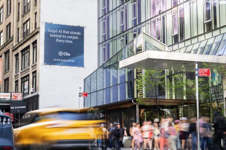 A Clio wallscape ad reading "Legal AI that stands behind its output. Every time." next to the AC Hotel Times Square as a yellow taxi blurs past.