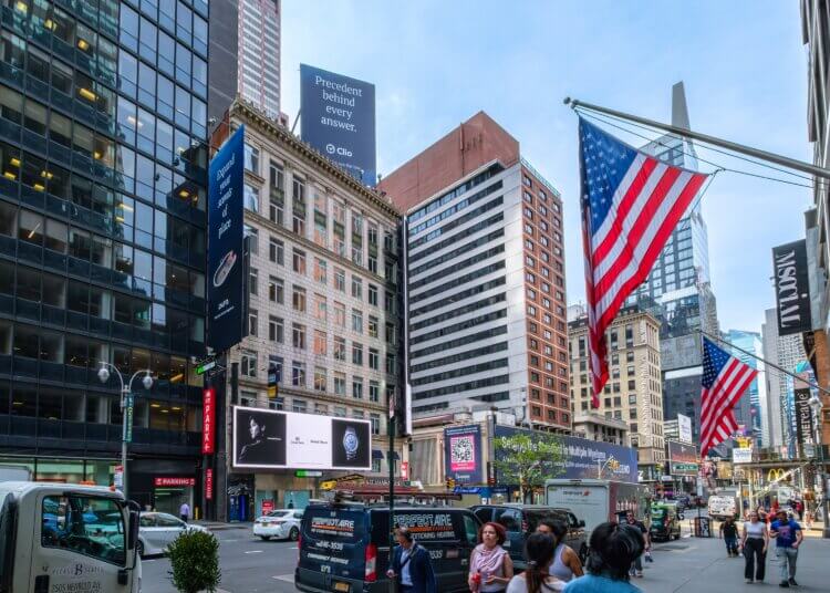 A rooftop Clio billboard reading "Precedent behind every answer." towering above buildings near Times Square with American flags in the foreground.
